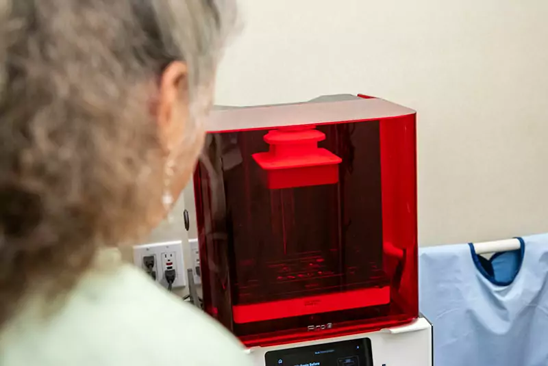 A person with gray hair, seen from behind, observes a red, transparent 3D printer in operation, conveying curiosity and technological engagement.