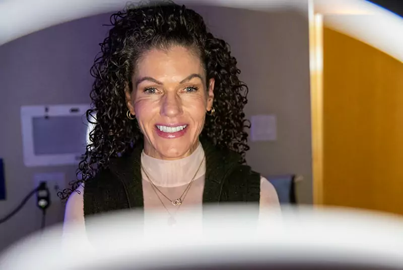 Smiling woman with curly hair, wearing a dark vest and light top. She appears in a softly lit room, framed by blurred elliptical shapes in the foreground.
