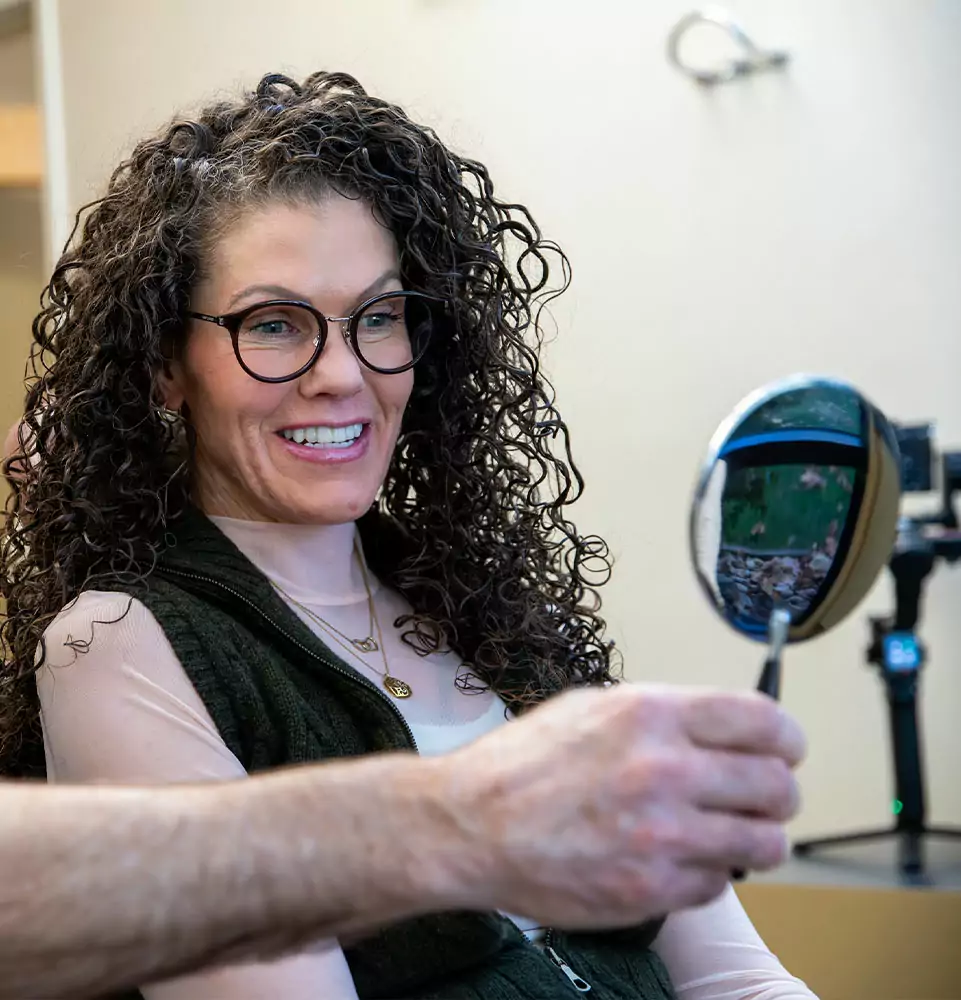 Woman with curly hair and glasses smiles while holding a small round mirror. A hand points at the mirror, suggesting a friendly consultation.