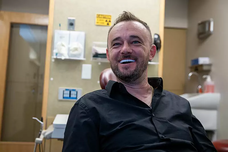 A man in a black shirt smiles while sitting in a dentist's office. The room has dental equipment and a radiation warning sign, creating a relaxed yet clinical atmosphere.