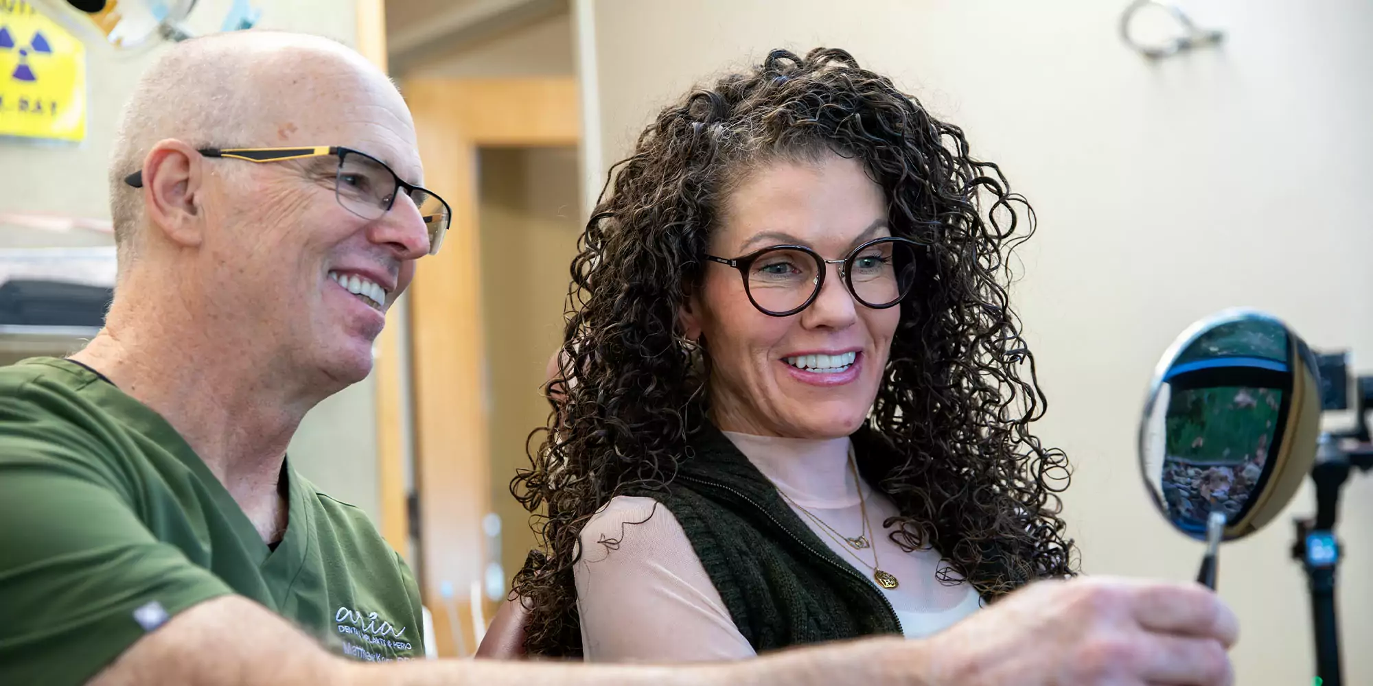 A man and woman smile while looking at themselves in a handheld mirror in an office. The atmosphere is friendly and relaxed, suggesting a positive interaction.