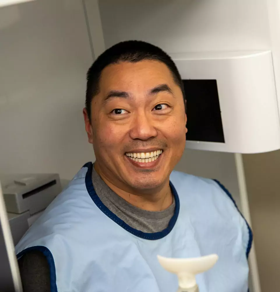 Smiling man in a blue protective apron in a dental or medical setting. He exudes happiness and ease, likely undergoing a procedure.