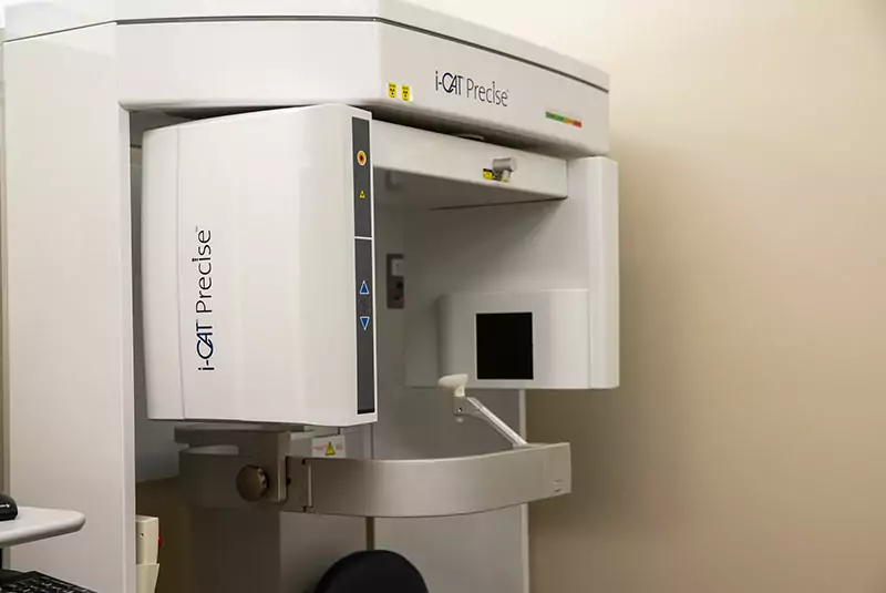 Dental imaging device in a medical room. The i-CAT Precise machine, with a sleek, white design, stands against a neutral-colored wall, conveying a clinical tone.