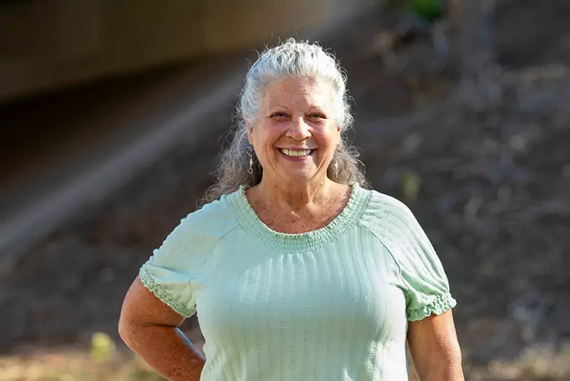 Smiling elderly woman with long gray hair in a light green blouse stands outdoors on a sunny day. Background shows blurred greenery and soil.