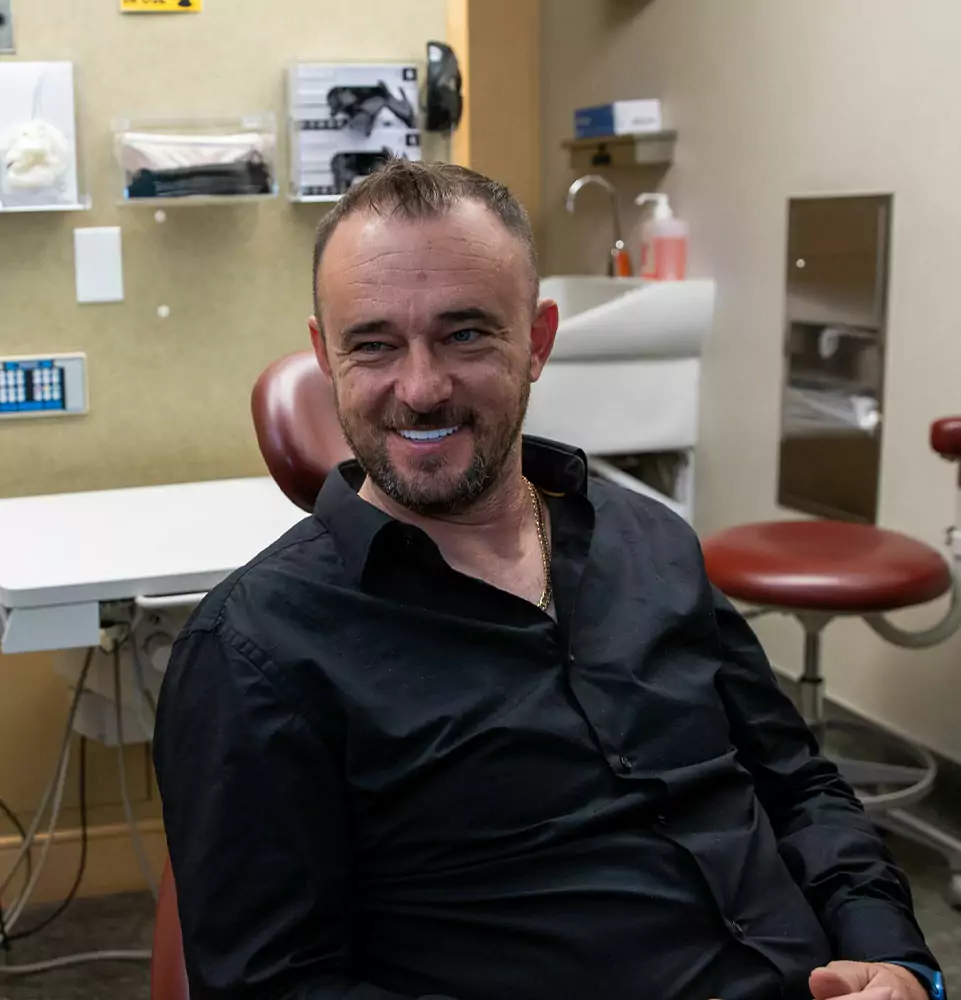 Man in a black shirt smiling while sitting in a well-lit dental office. Background includes dental equipment and chairs, creating a relaxed atmosphere.