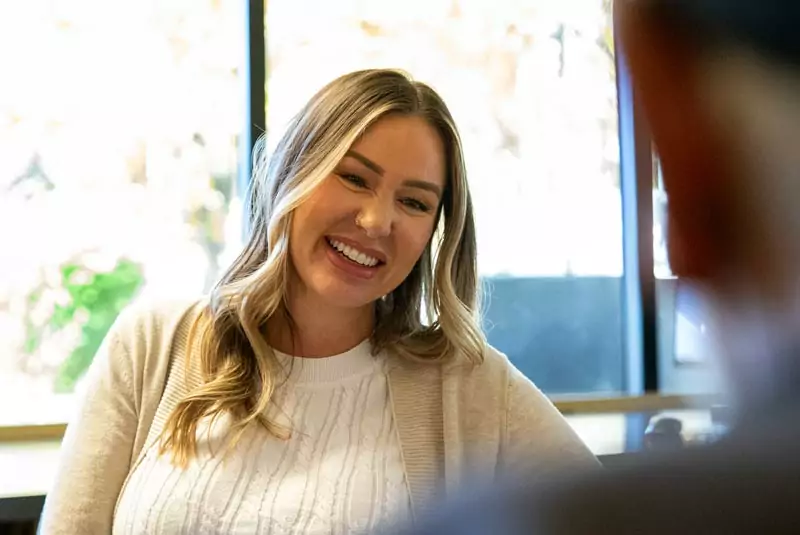 A smiling man in a black shirt sits against a neutral beige background, conveying a warm and welcoming demeanor. The background is softly blurred.