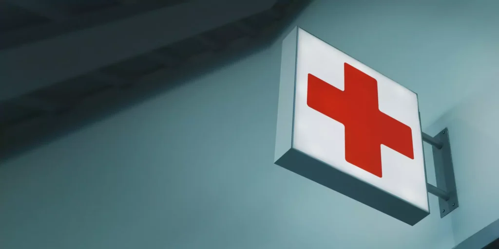 A wall-mounted, illuminated sign displays a large red cross against a white background, symbolizing medical or emergency services. The setting has a calm, sterile atmosphere.