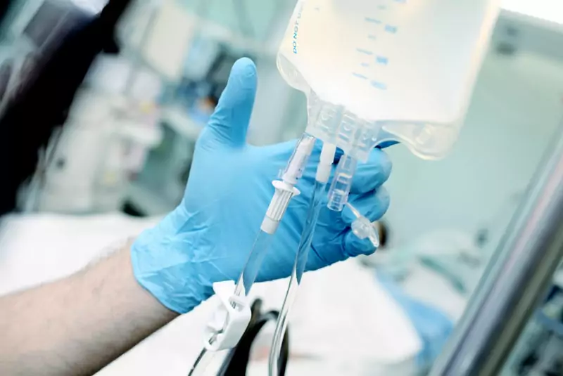 A gloved hand adjusts an IV drip in a medical setting, conveying precision and care. The blurred background suggests a hospital environment.