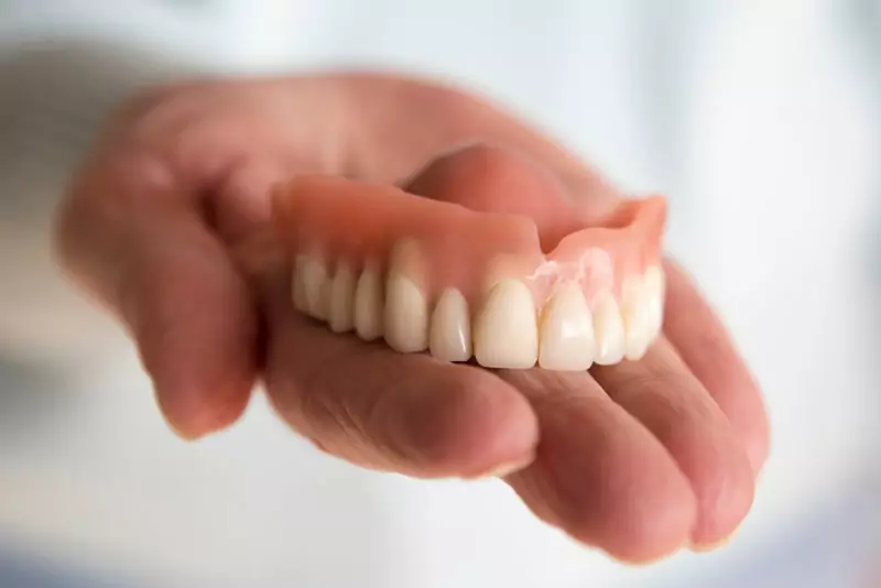 A close-up of a hand holding a set of upper dentures with pink gums and white teeth, against a soft, blurred background, conveying care and precision.