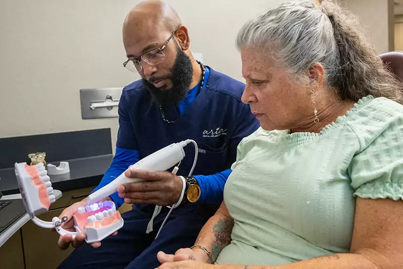 A dentist demonstrates a dental scanner to a seated woman, holding a model of teeth. The setting is a dental office, and their expressions are attentive.