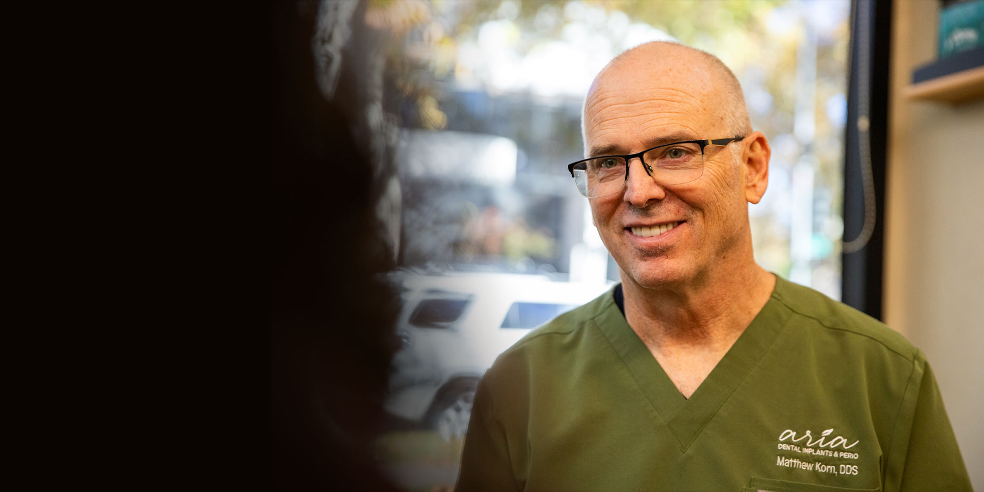 A man in green scrubs, wearing glasses, smiles warmly. He's indoors by a window with blurred trees outside, creating a friendly, welcoming tone.