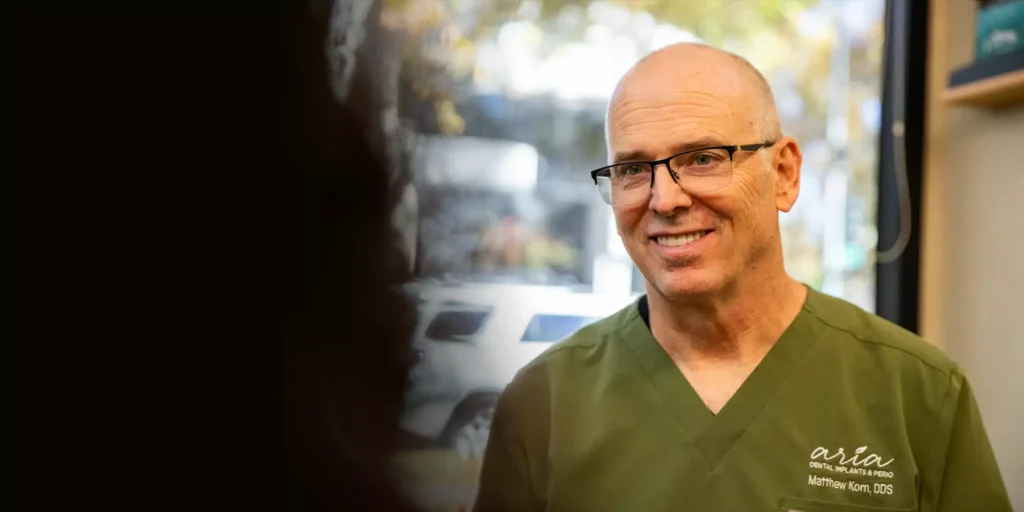 A smiling man in glasses and green scrubs stands indoors, with his name embroidered on his shirt. A blurred window with trees visible behind him.