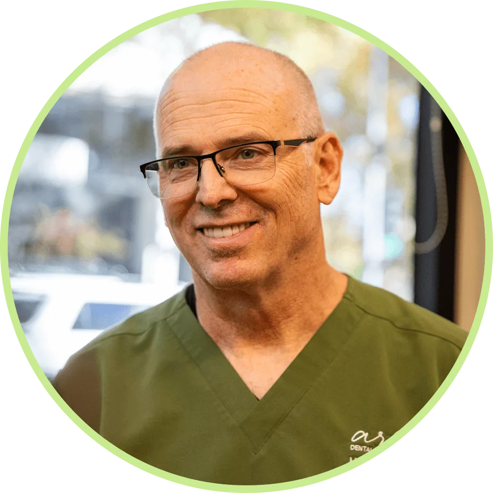 Man in green scrubs and glasses smiling warmly, seated indoors against a blurred background. The mood is professional and approachable.