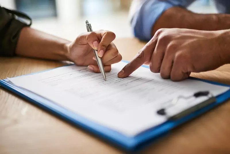 Close-up of two hands reviewing a document on a clipboard. One hand holds a pen poised to write, while the other points, indicating collaboration.