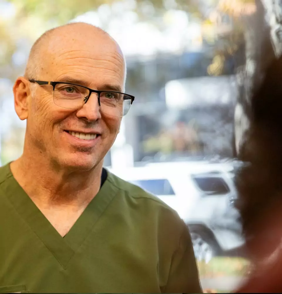 Bald man with glasses in green scrubs smiling warmly indoors, conveying friendliness. Blurred background with a hint of a white SUV outside.