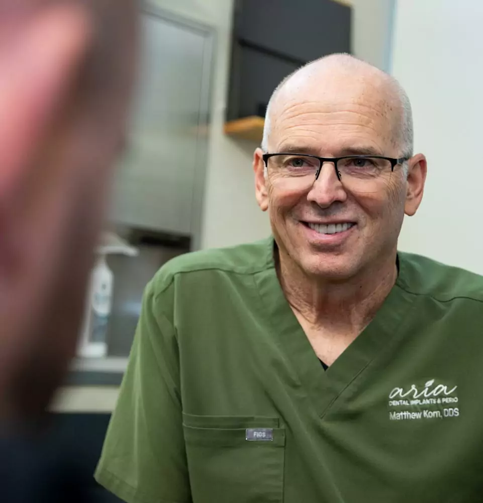 A smiling dentist in green scrubs listens attentively, conveying friendliness and professionalism. The setting appears to be a dental office.
