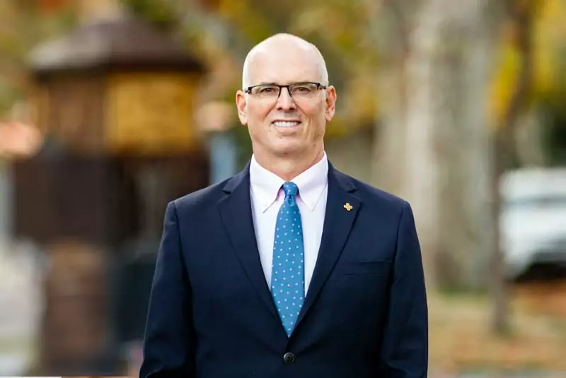 A man in a suit with a blue polka dot tie stands outdoors. The background is blurred with earthy tones, suggesting a fall setting. He looks content and professional.