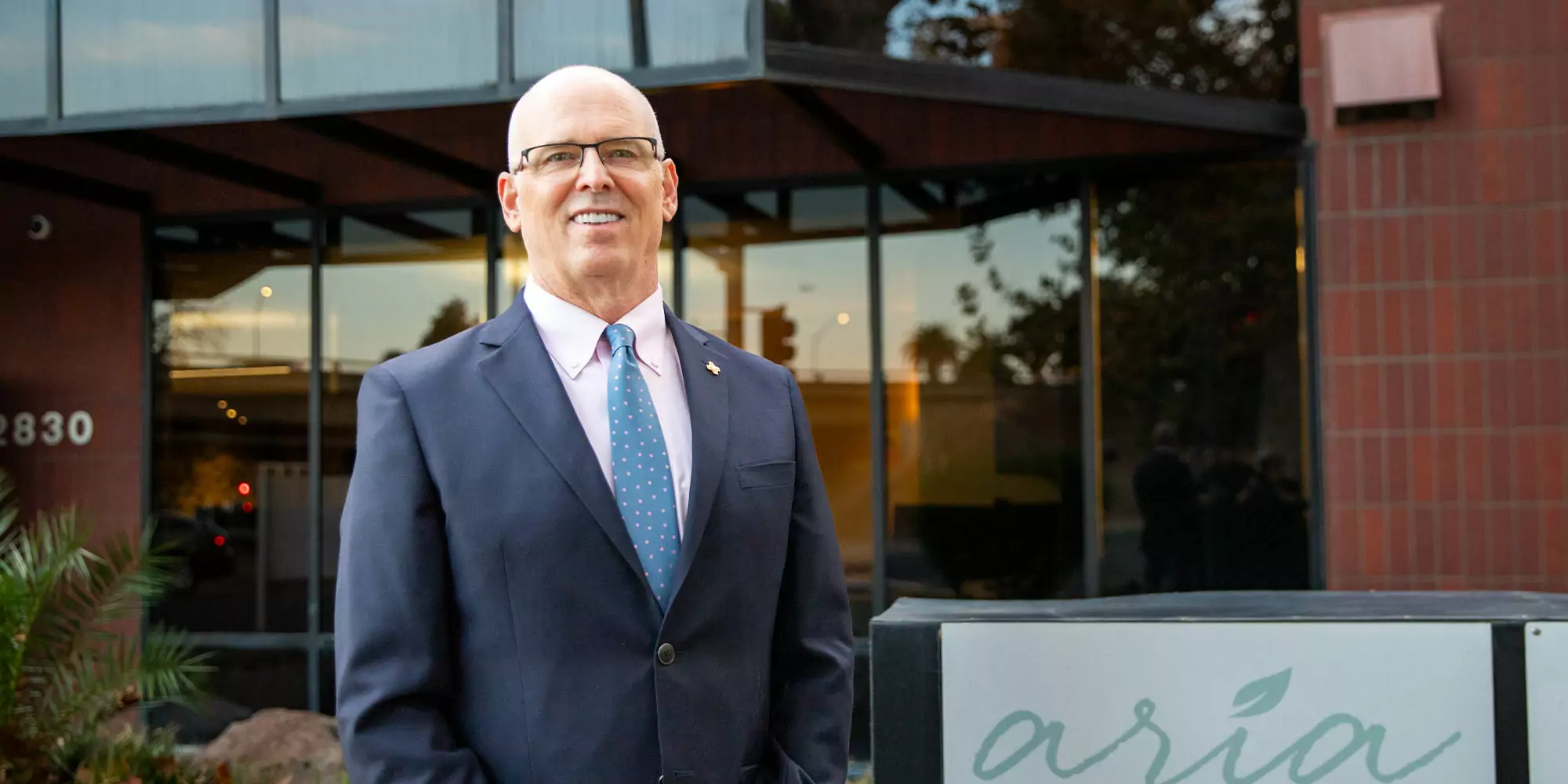 A man in a suit and tie smiles confidently outside a modern glass building with "aria" on a sign. The setting is professional and welcoming.