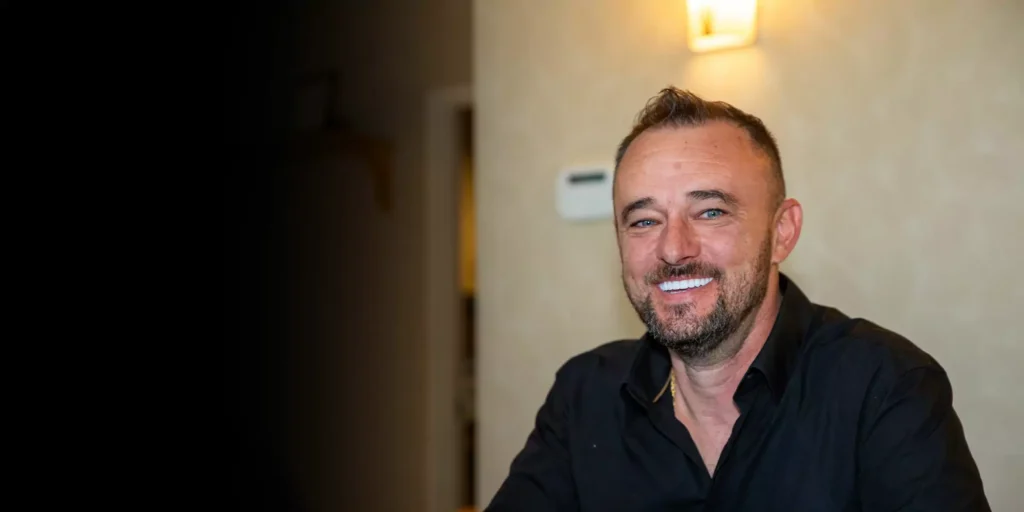 Smiling man with short hair and beard, wearing a black shirt, sits indoors against a beige wall with a warm light above, exuding a friendly tone.