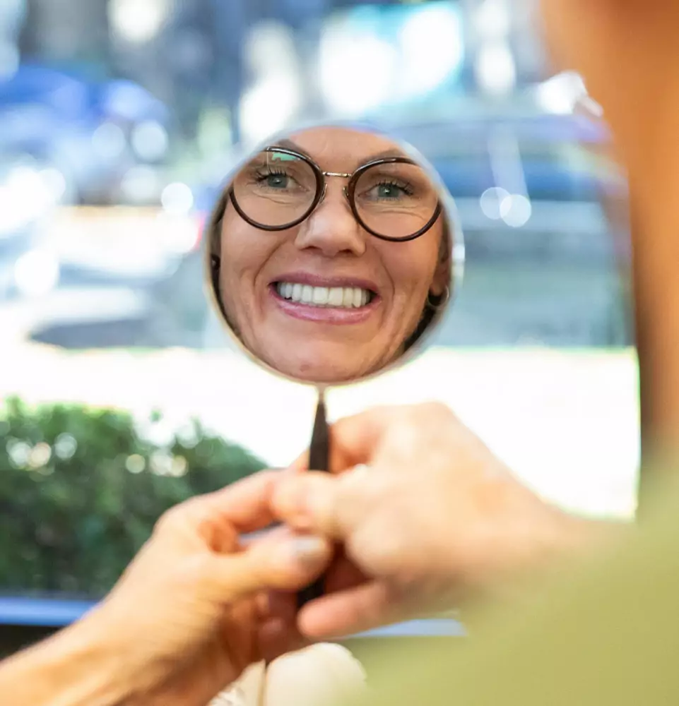 A woman with glasses smiles warmly at her reflection in a handheld mirror. The background is softly blurred, creating a cheerful and relaxed atmosphere.
