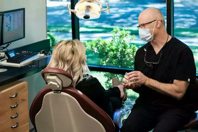 A dentist in a mask discusses dental models with a patient in a chair, set against a window view of greenery. The scene feels professional and calm.
