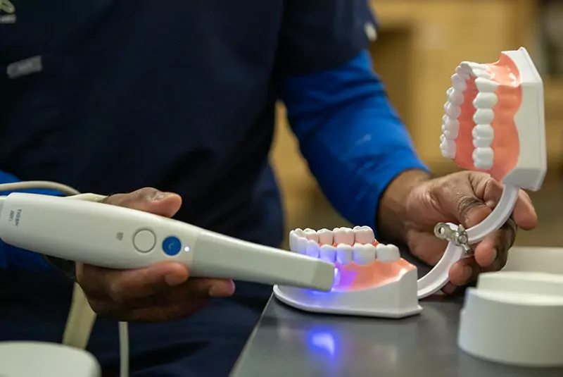 A dentist uses a dental scanner on a model of teeth, emitting a blue light. The setting is clinical, with a focused and professional tone.
