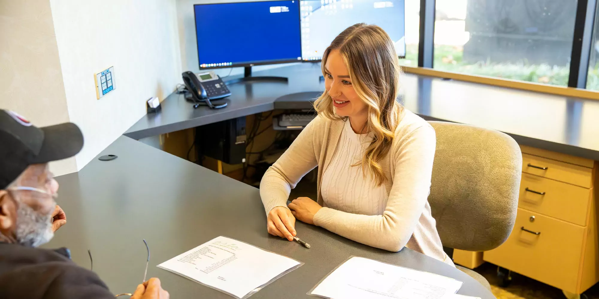 A woman with long hair smiles warmly at a man during a meeting at an office desk. Papers and pens are on the table, and computers are in the background.