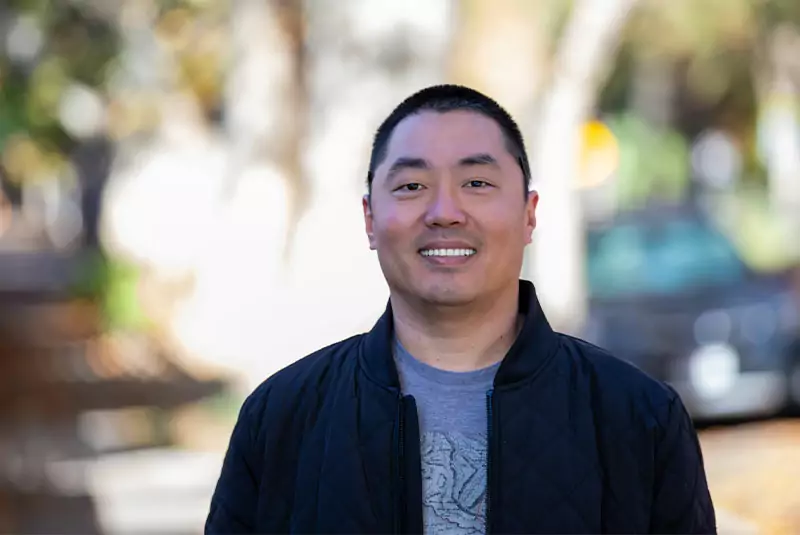 A smiling man in a black jacket stands outdoors with a blurred, sunny background of trees and a car, conveying a relaxed and cheerful mood.