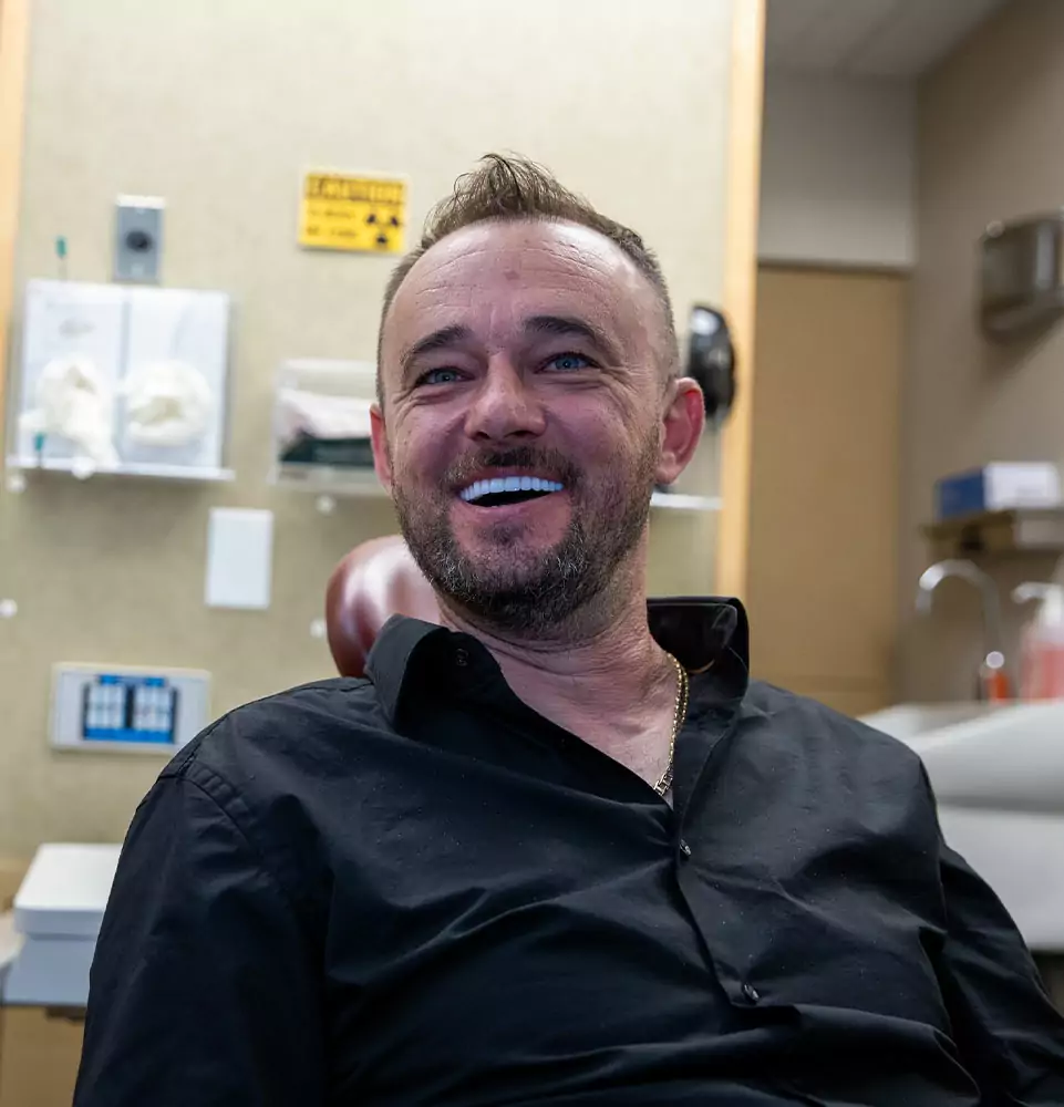 A smiling man with a beard sits in a clinic room wearing a black shirt. The environment is relaxed with medical supplies in the background.