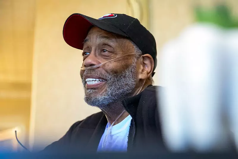 Elderly man wearing a black cap and a nasal oxygen tube, smiling warmly. Soft lighting, blurred foreground and background, conveying a serene mood.