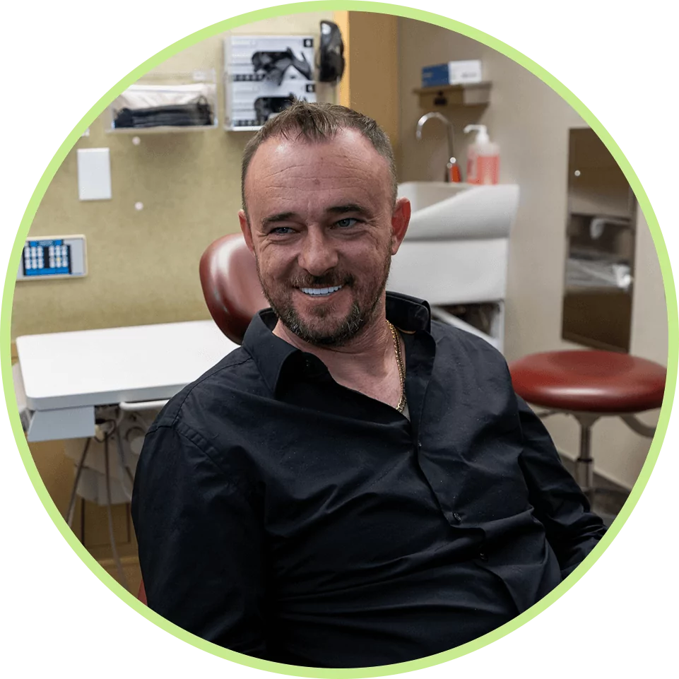 Smiling man wearing a black shirt seated in a medical office. Background shows medical supplies and equipment, creating a calm, professional atmosphere.