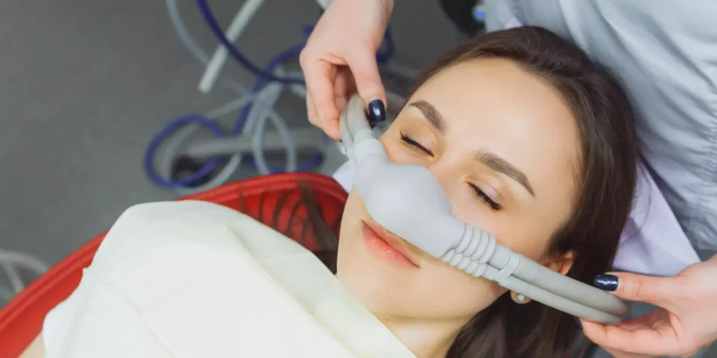 A woman reclines with eyes closed, wearing a nasal mask in a medical setting. Hands adjust the mask, conveying calm and care. Mixed emotions are present.