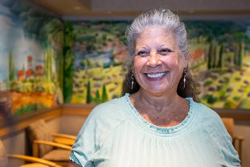 Smiling woman with gray hair in a light green shirt in front of a colorful mural depicting a scenic countryside. The atmosphere is warm and joyful.