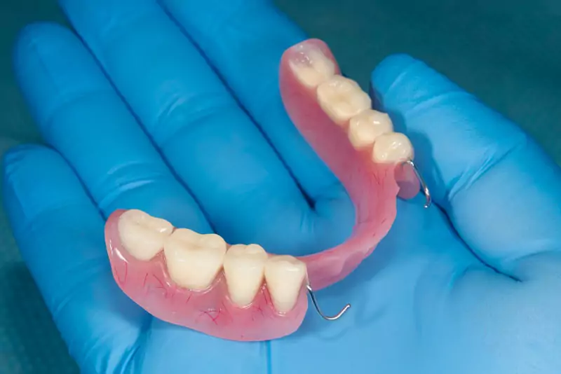 A gloved hand holds a partial denture with pink gums and white teeth, showcasing dental care tools. The blue glove contrasts with the realistic prosthetic.