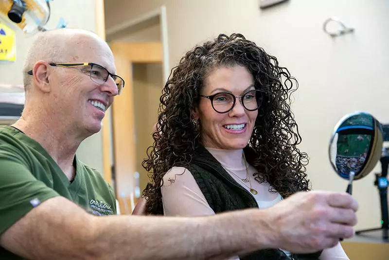 A woman with curly hair smiles into a small mirror held by a bald man with glasses in a green shirt. They appear happy and engaged in a friendly setting.