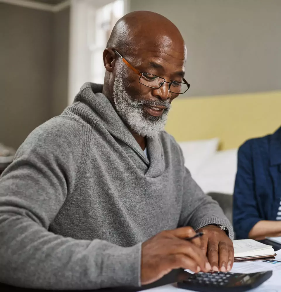 A man with glasses and a beard sits at a table, smiling softly while using a calculator. He's wearing a gray sweater, evoking a calm and focused mood.