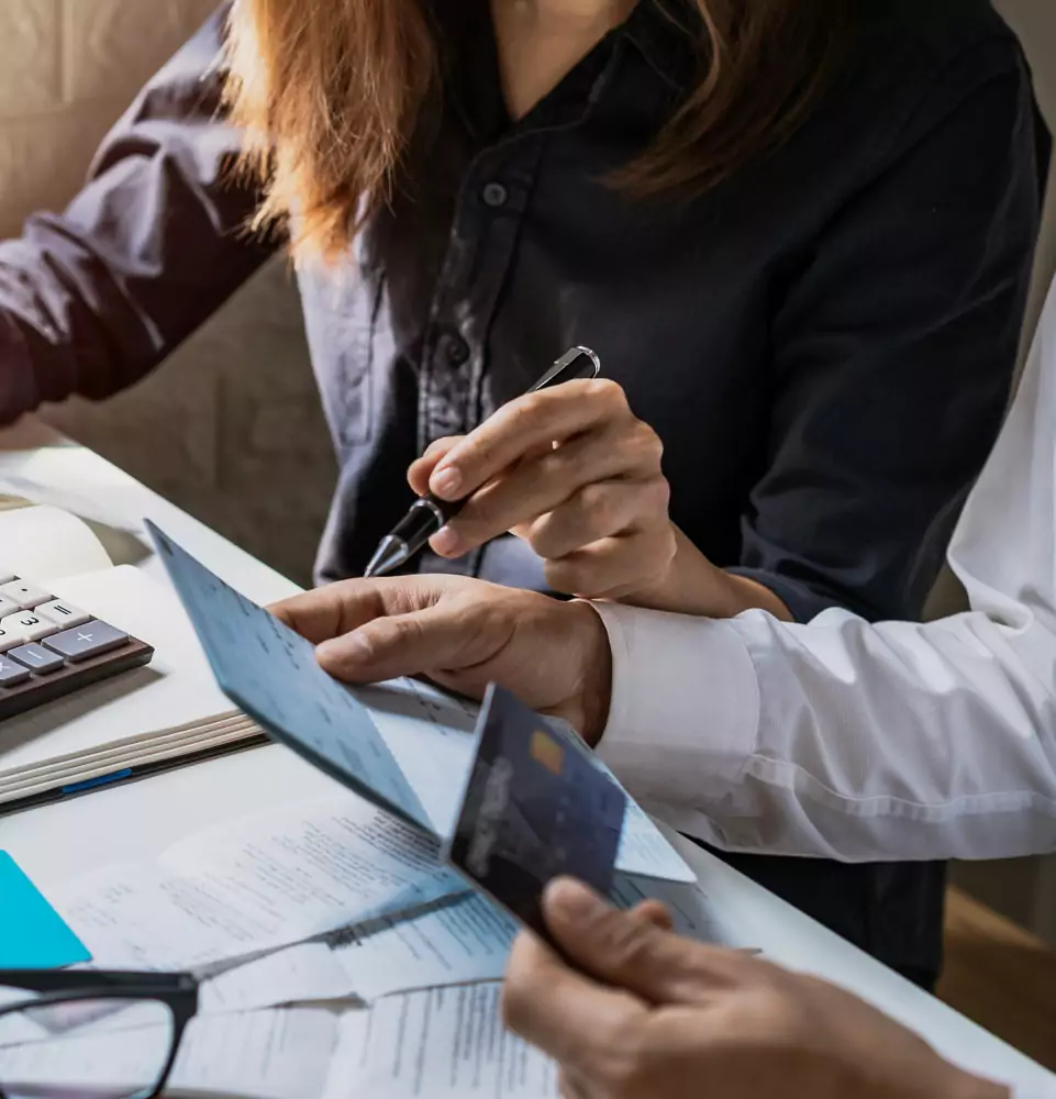 Two people are reviewing financial documents at a desk. One points with a pen at a paper, while the other holds a credit card. A calculator and receipts are visible.