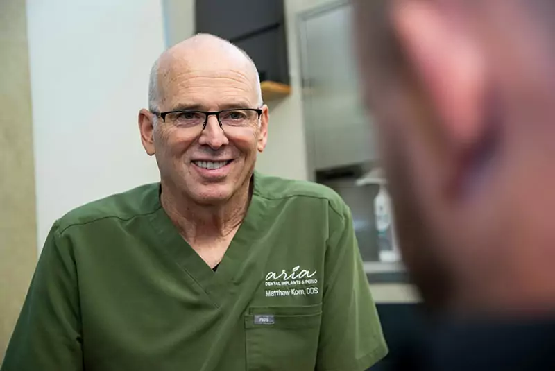 Smiling dentist in green scrubs with glasses speaks to a patient, conveying warmth and professionalism. Dental equipment is visible in the background.