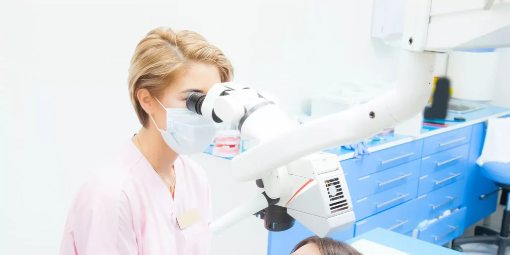 A dentist in a pink uniform and mask examines a patient using a dental microscope in a bright clinic. The setting feels professional and focused.