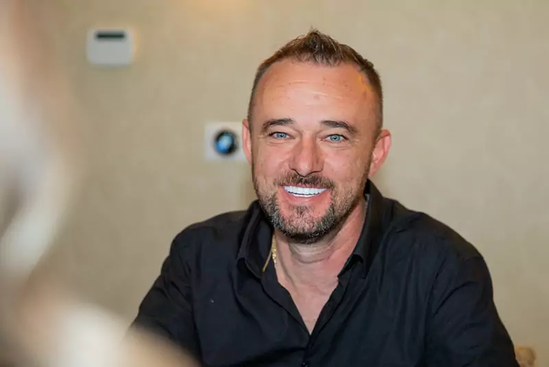Smiling man with short hair and beard in a black shirt sits indoors. Neutral background with soft lighting conveys a warm, friendly tone.