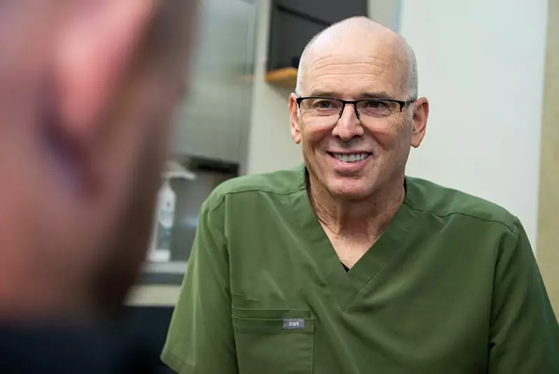 A person in green medical scrubs, wearing glasses, is smiling warmly during a conversation with another individual. The setting suggests a healthcare environment.