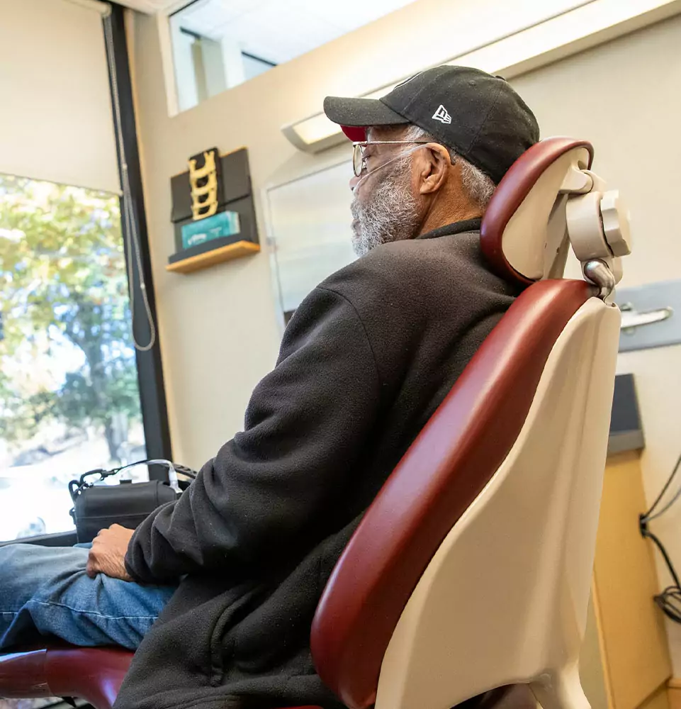 An elderly man wearing a black cap and jacket sits in a dental chair, gazing out a window. The setting is calm and clinical, with soft lighting.