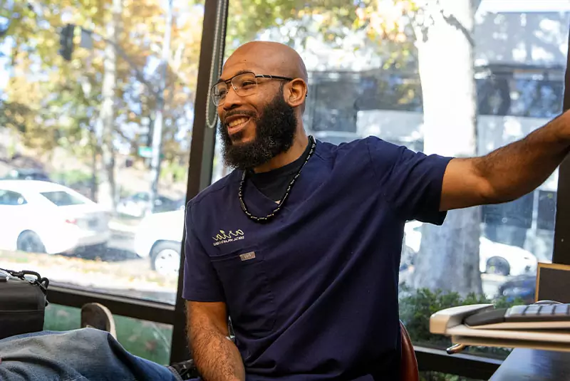 A smiling man in navy scrubs sits near a large window, gesturing with his arm. Sunlight filters through autumn trees outside, creating a warm atmosphere.