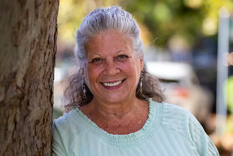 Smiling older woman with long gray hair and earrings leans against a tree outdoors. She wears a light green top. Background shows blurred greenery.