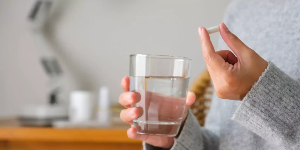 A person in a gray sweater holds a pill in one hand and a glass of water in the other, suggesting a moment of taking medication. The background is softly blurred.