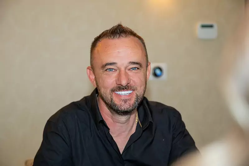 Smiling man with short hair and a beard, wearing a black shirt, sits indoors against a light-colored wall. The atmosphere is warm and friendly.