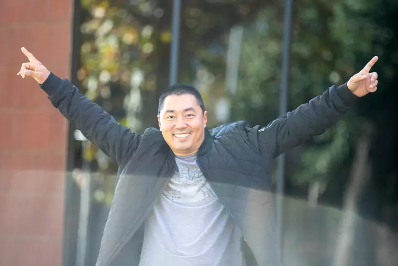 A man in a black jacket joyfully raises both arms in a celebratory gesture. He smiles widely against a blurred outdoor background, conveying happiness.