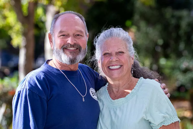 A smiling older couple poses together outdoors. The man in a blue shirt and the woman in a light top, both appear happy and relaxed with blurred greenery in the background.