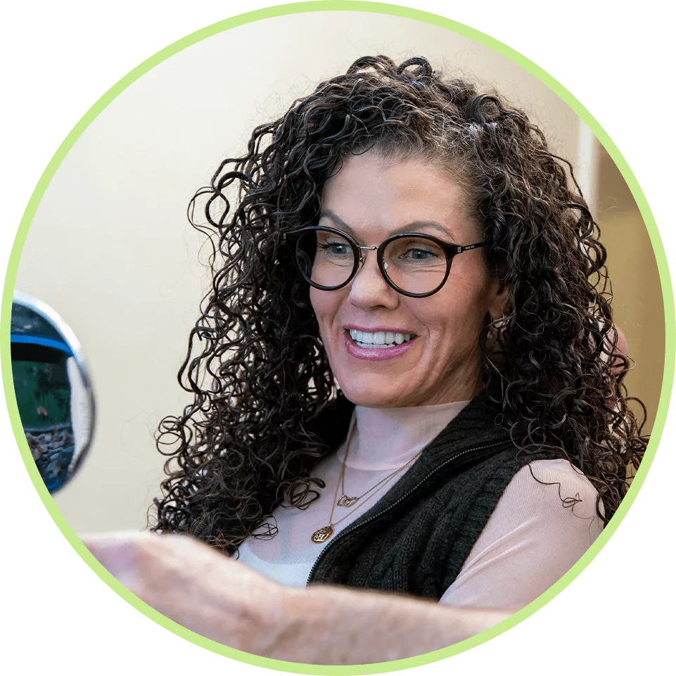A smiling woman with curly hair and glasses holds a mirror, wearing a pink top and black vest. The background is softly blurred, conveying a joyful atmosphere.