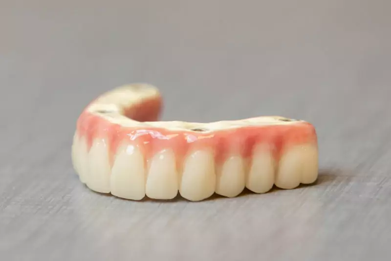 A close-up of an upper dental denture on a gray surface, showing realistic teeth and pink gums. The tone is clinical and neutral.